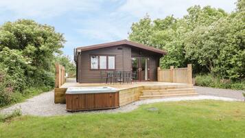 Eine kleine, eingeschossige Holzhütte mit Whirlpool und Sitzgelegenheit im Freien, umgeben von einer Holzterrasse und einem Kiesweg.