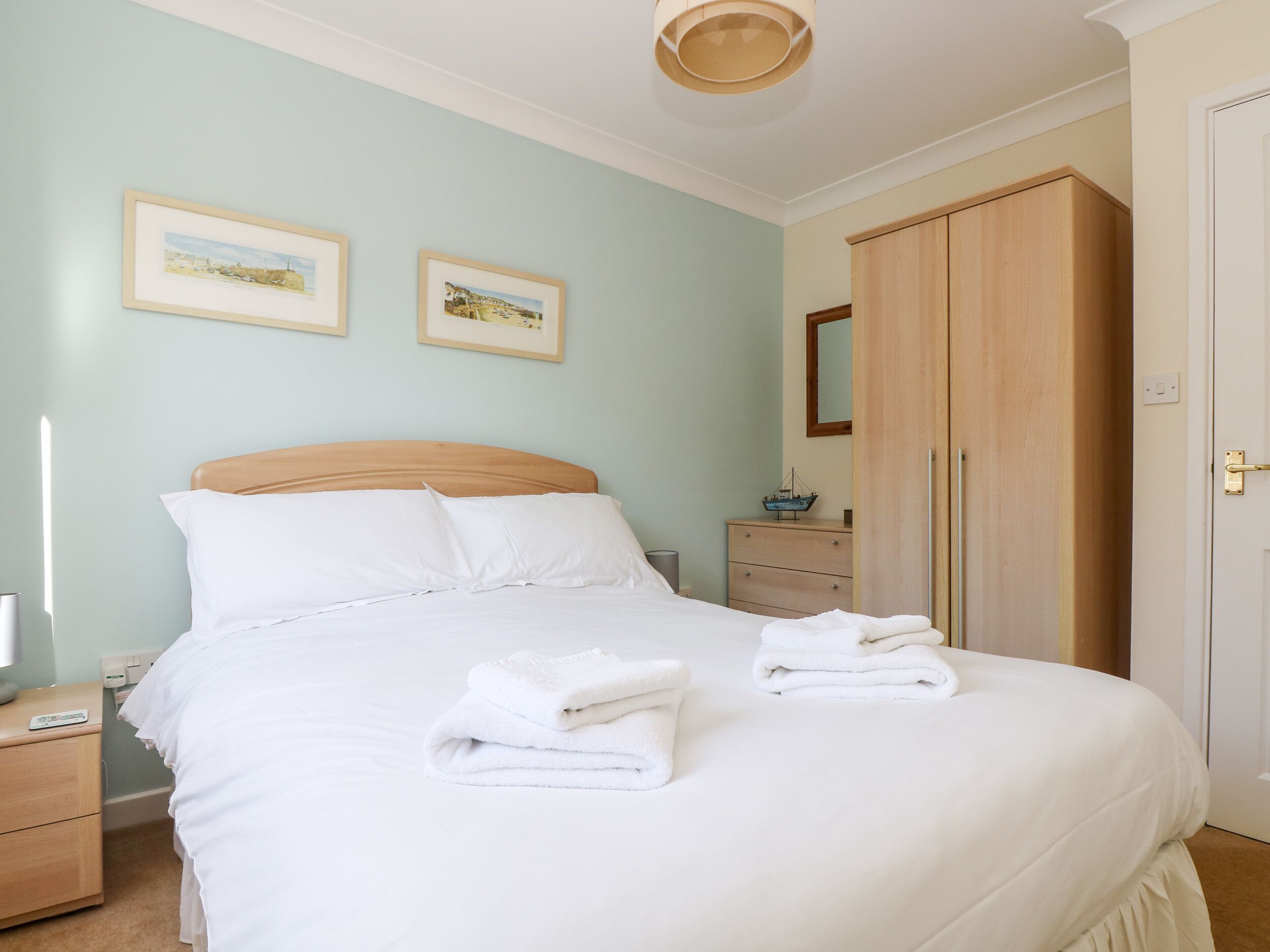 A neatly made bed with white linens and towels, a wooden headboard, a nightstand with a lamp, a dresser, and two framed pictures on the wall.