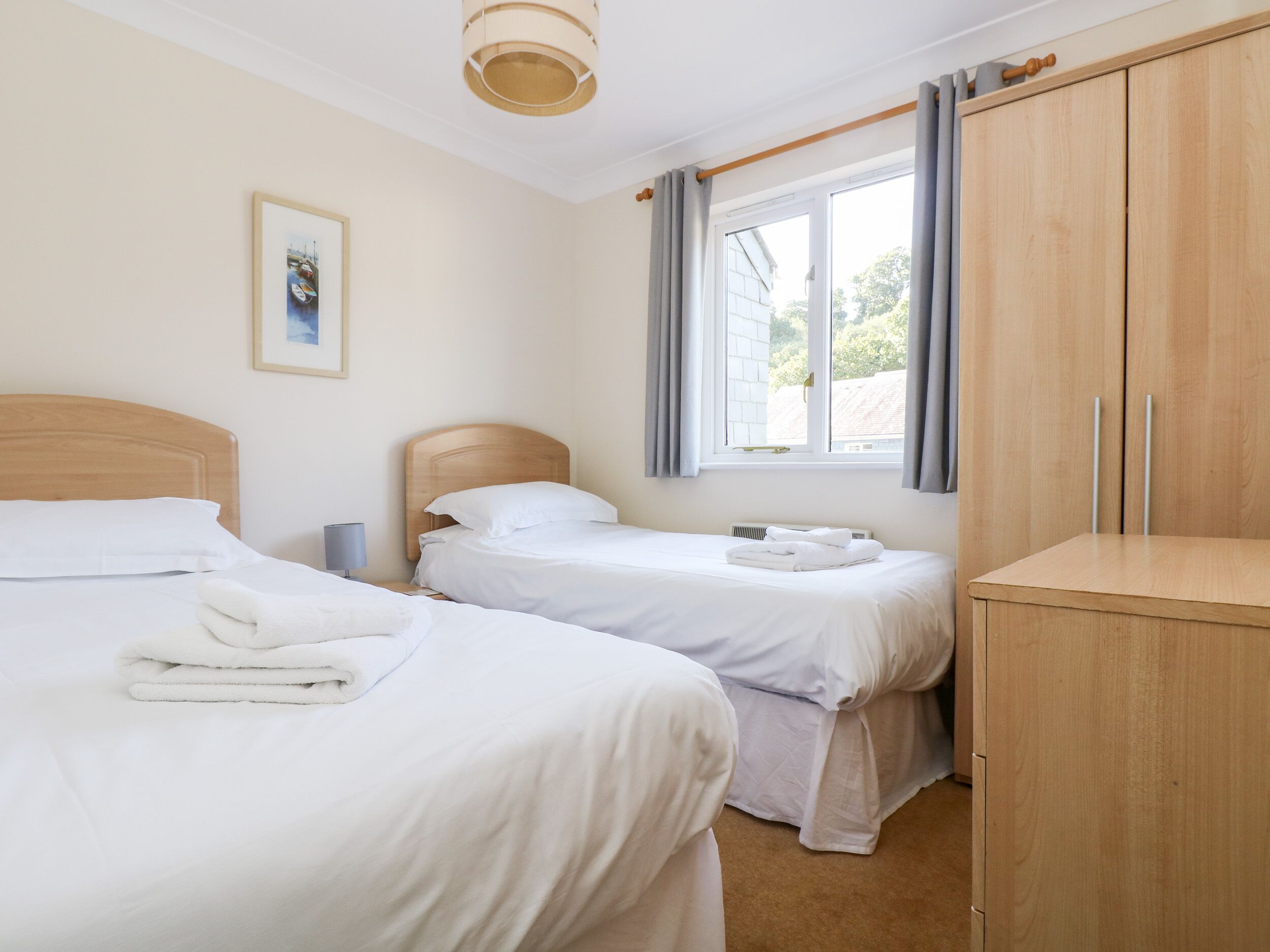 A hotel room with two single beds, a wooden wardrobe, a window with curtains, and a framed picture on the wall.