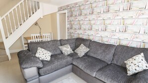 Ein graues Ecksofa mit gemusterten Kissen, eine weiße Treppe und ein Bücherregal mit Büchern und Dekoartikeln.