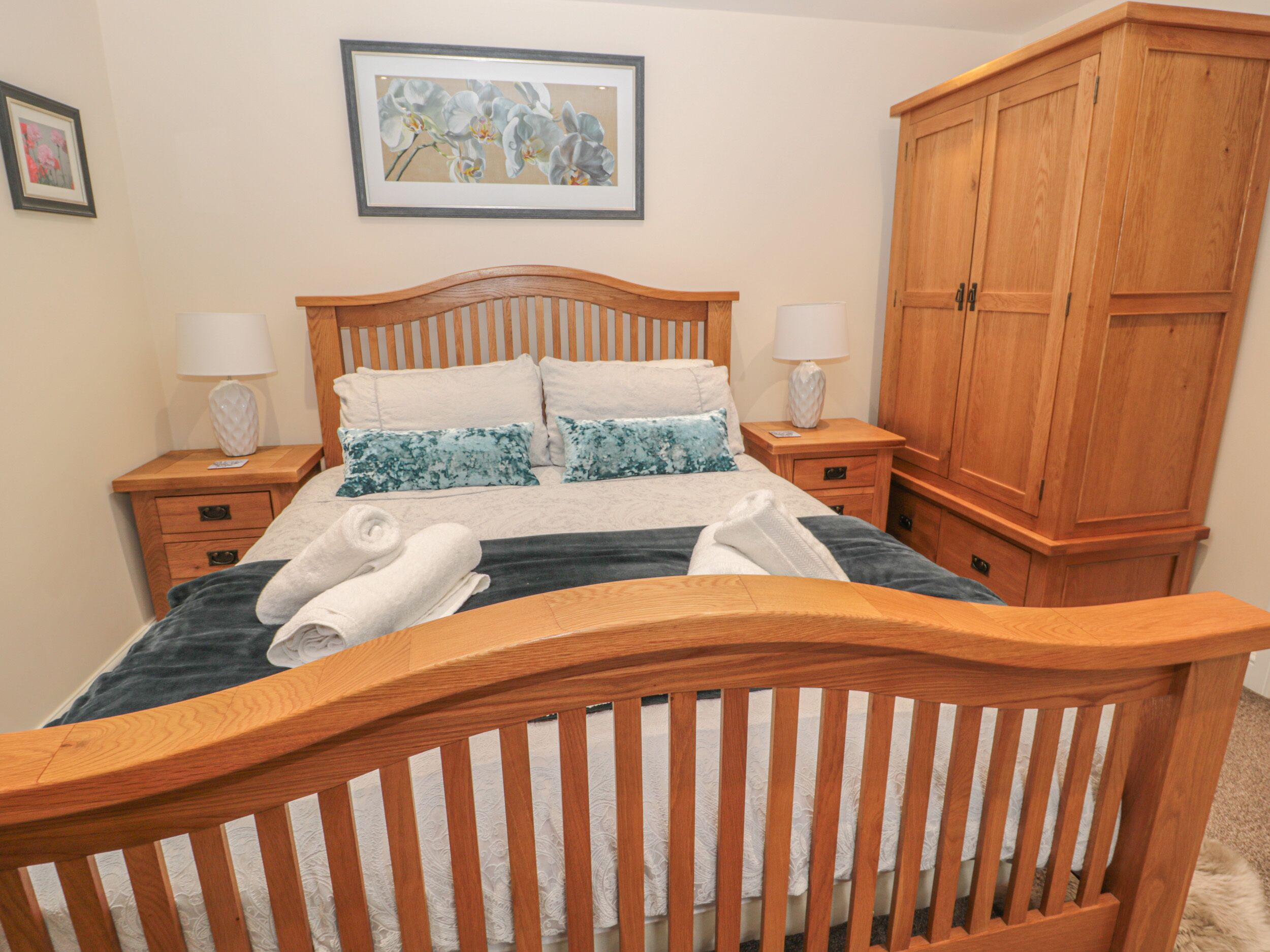 A bedroom with a wooden bed, two bedside tables with lamps, a wooden wardrobe, and a framed floral artwork on the wall.