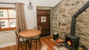 A cozy room with a stone wall, a wooden table with chairs, a window with curtains, and a wood-burning stove. - Heugh Barn (Richmond)