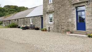 A stone building with a gravel driveway, a blue door, and a small outdoor seating area with a table and chairs. - Seaview-barsloisnach Cottage (Lochgilphead)