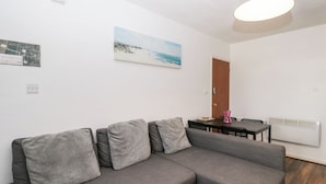 A modern living room with a grey sectional sofa, a dining table with chairs, a painting of a beach, and a ceiling light fixture. - Ocean Breeze (Burnham-on-Sea)