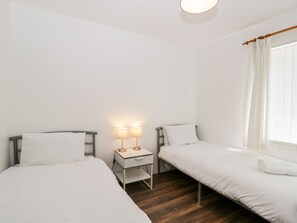 A room with two single beds, a small bedside table with lamps, a wooden floor, and a window with curtains.
