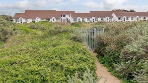A row of white houses with brown roofs, surrounded by dense greenery and a sandy path leading to a gate. - Ocean Breeze (Burnham-on-Sea)
