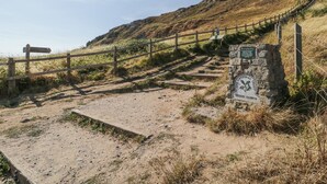 A dirt path leading to a stone sign with a map, a wooden fence, and a grassy hill in the background. - Ocean Breeze (Burnham-on-Sea)