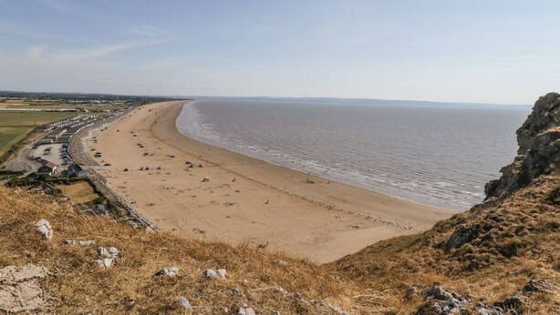 A coastal beach with a long stretch of sandy shore, a rocky cliff on the right, and a road running parallel to the beach. - Ocean Breeze (Burnham-on-Sea)