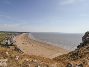 A coastal beach with a long stretch of sandy shore, a rocky cliff on the right, and a road running parallel to the beach. - Ocean Breeze (Burnham-on-Sea)