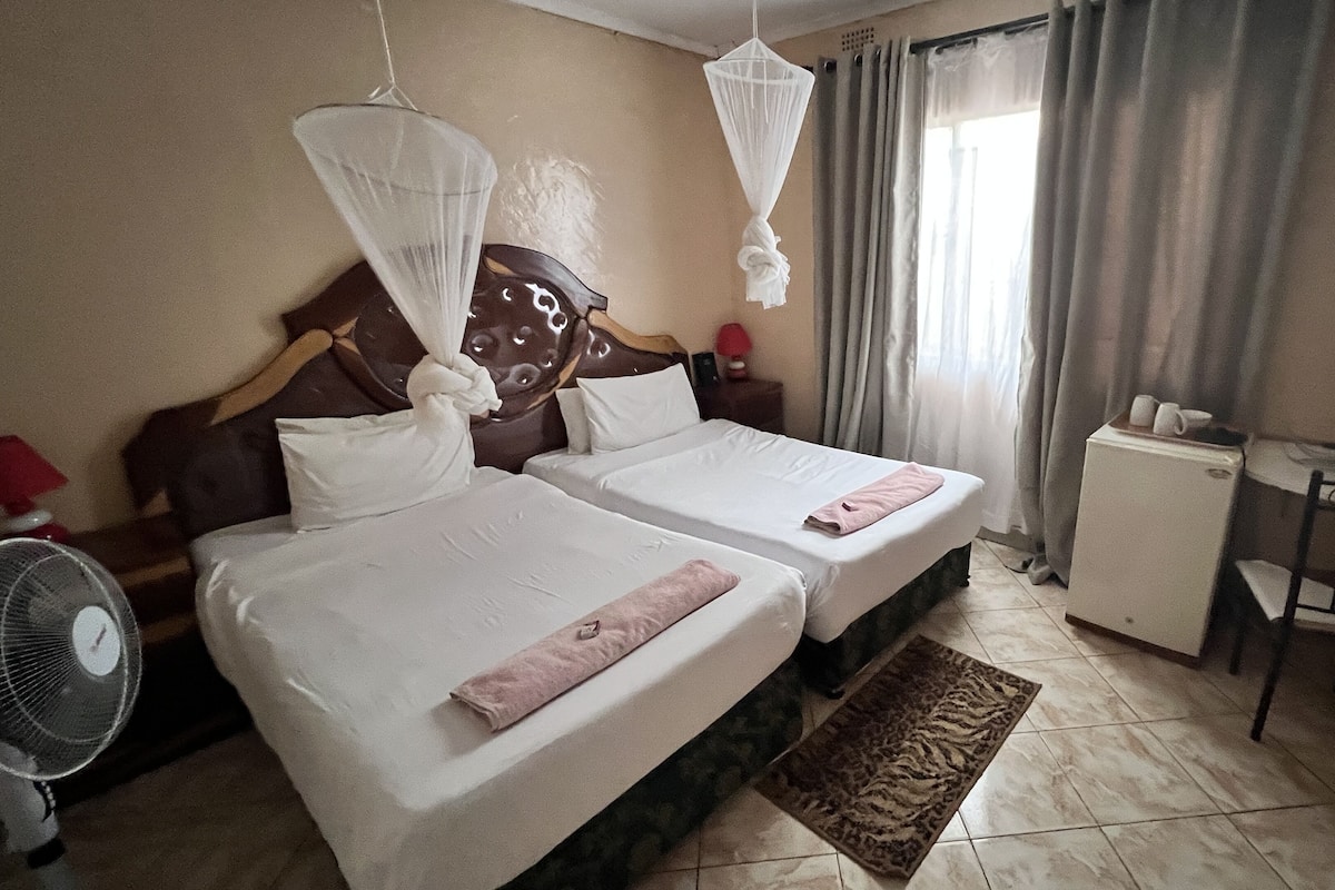 A hotel room with two beds, a bedside table, a chair, a fan, and a small heater.