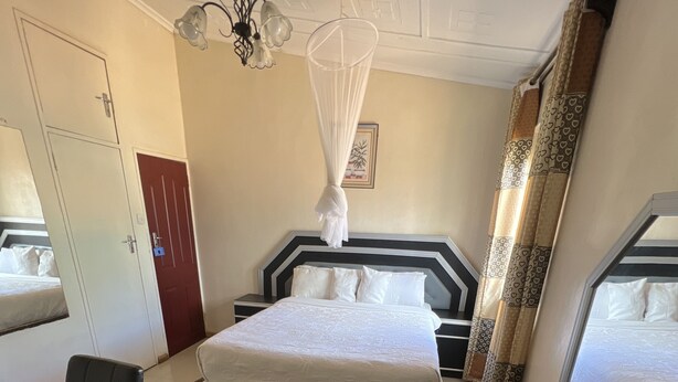A hotel room with a large bed, a desk with a kettle and mug, a chair, and a bedside table. - Nalikwanda Guest House (Kabwe)