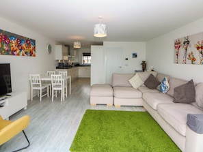 A modern living room with a sofa, a dining table with chairs, a TV, and a kitchen in the background.