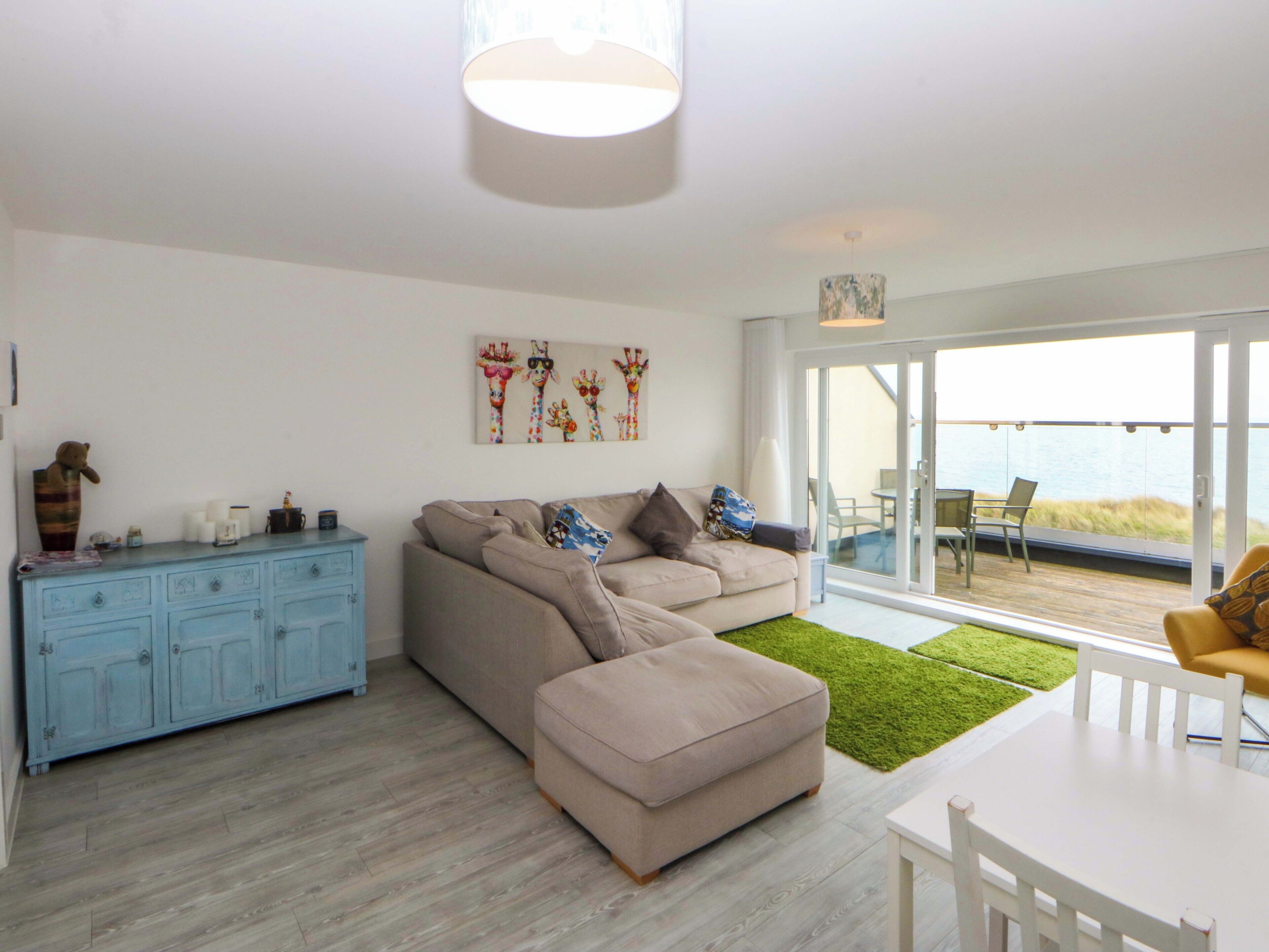 A modern living room with a sofa, a dining table, and a view of the sea through large glass doors.