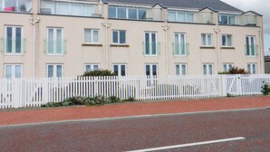 A multi-story residential building with a flat roof, white picket fence, and a paved road in front.