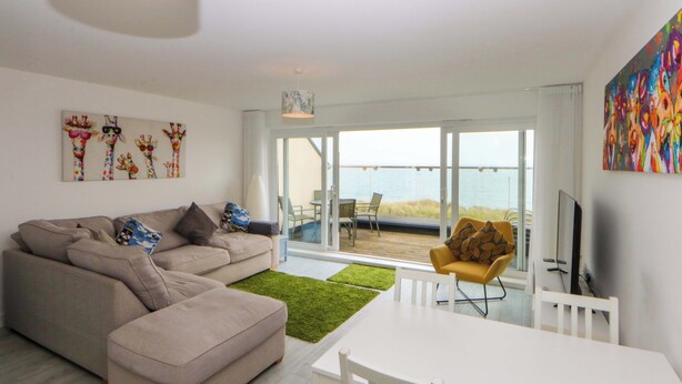 A modern living room with a sofa, a dining table, and a view of the sea through large glass doors.