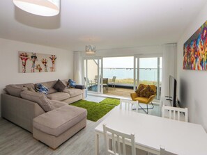 A modern living room with a sofa, a dining table, and a view of the sea through large glass doors. - Welymora (Pwllheli)
