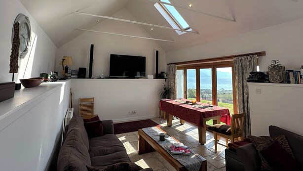 A spacious living area with a high ceiling, a flat-screen TV, a dining table, and a view through large windows. - Solas (Sleat)