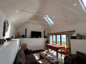 A spacious living area with a high ceiling, a flat-screen TV, a dining table, and a view through large windows. - Solas (Sleat)
