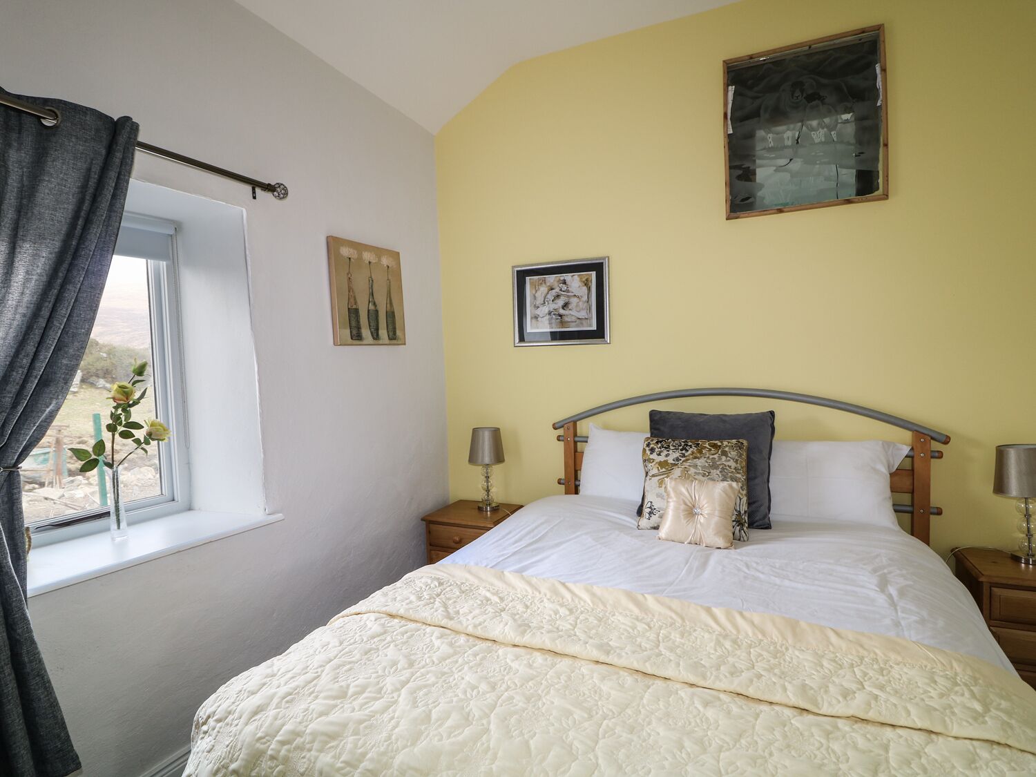 Un dormitorio con cama, una ventana con cortinas, una mesita de noche con lámpara y dos cuadros colgados en la pared.