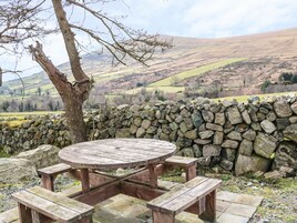 Una mesa de picnic de madera rústica con bancos, frente a un muro de piedra y un paisaje de colinas onduladas.