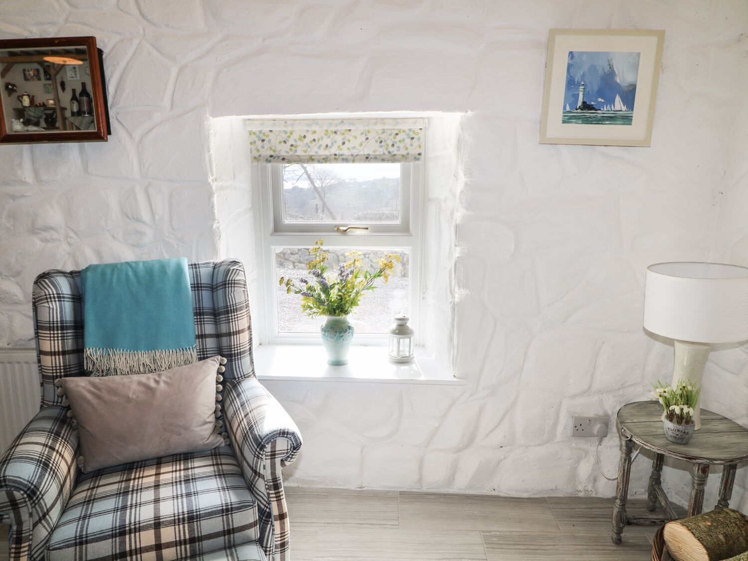 Una habitación acogedora con un sillón a cuadros, una mesita con lámpara, una ventana con vistas y un cuadro en la pared.