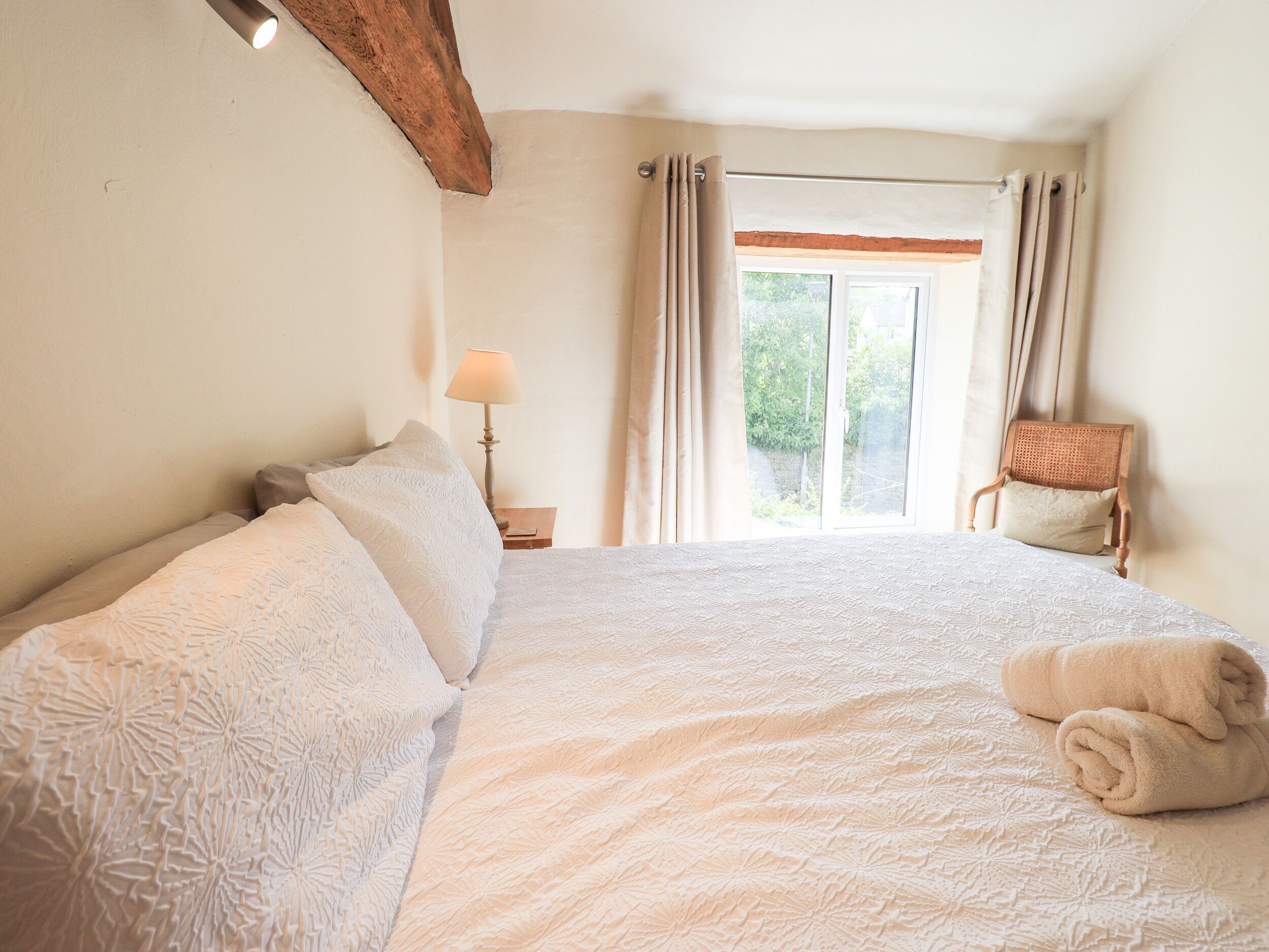 Un dormitorio con una cama grande, una mesita de noche, una lámpara, una silla y una ventana con cortinas.
