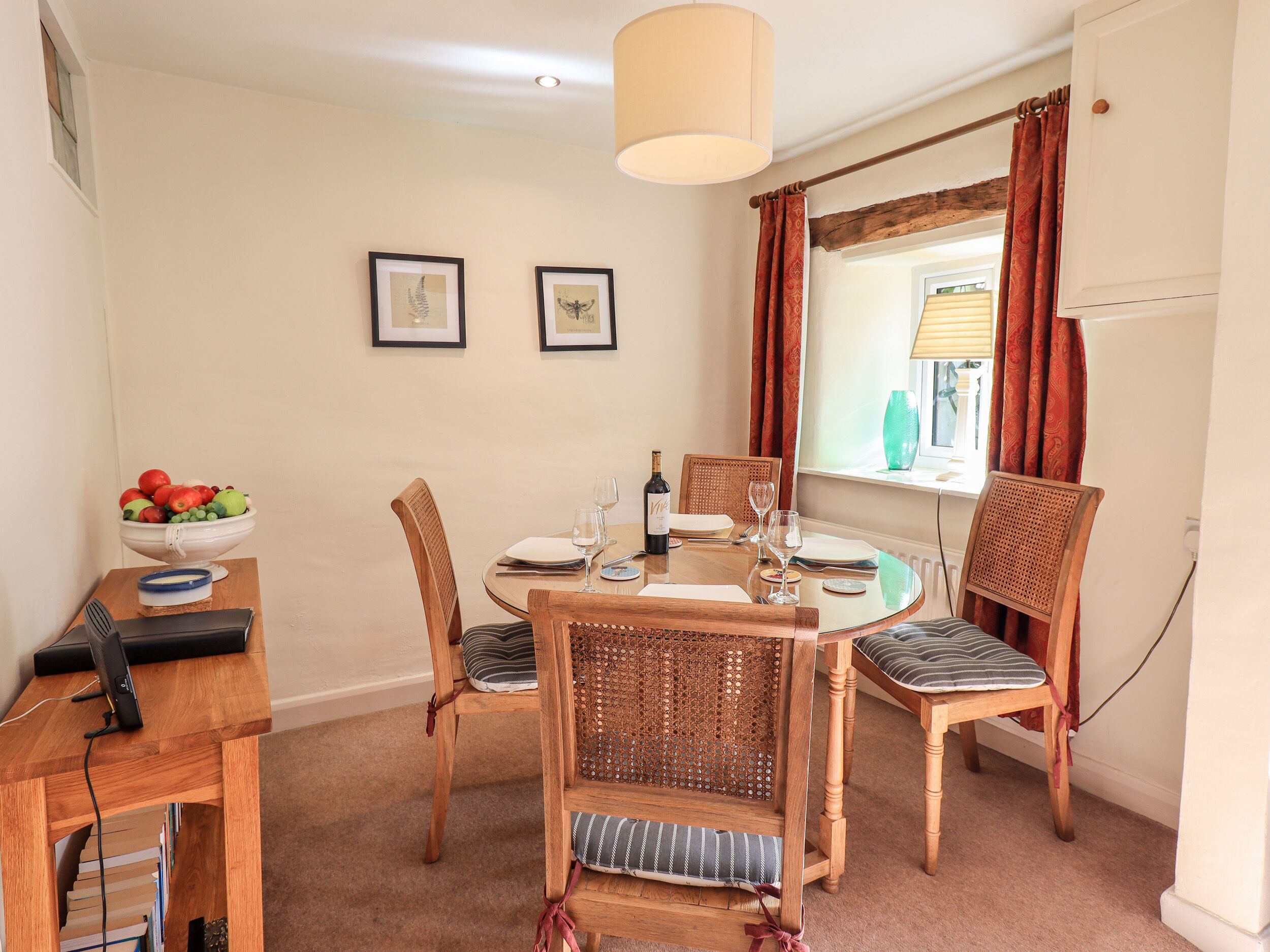 A dining area with a round table, four wooden chairs, a bowl of fruit, a bottle of wine, and two framed pictures on the wall.