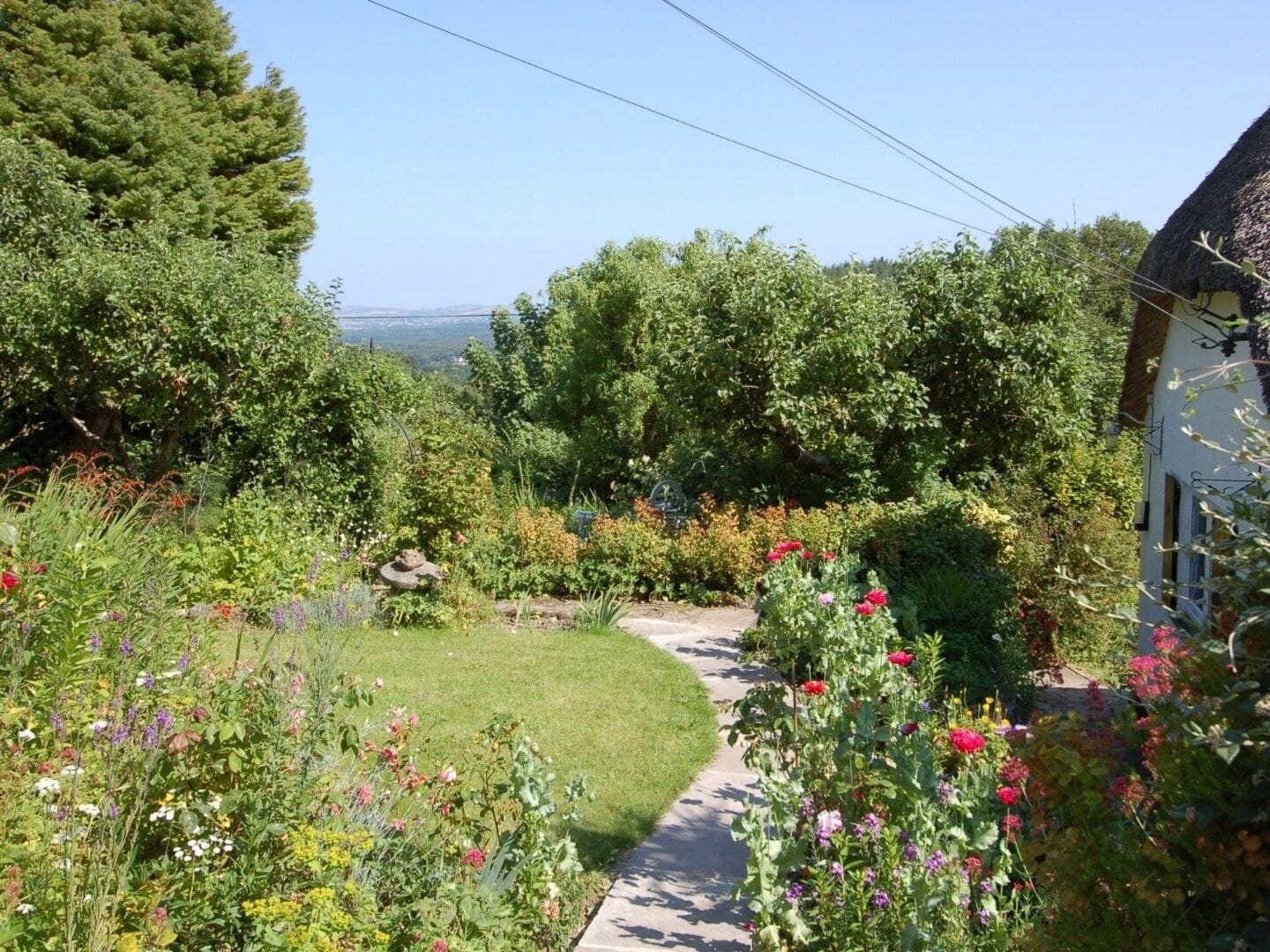 A garden pathway leading to a house with a thatched roof, surrounded by lush greenery and flowering plants.