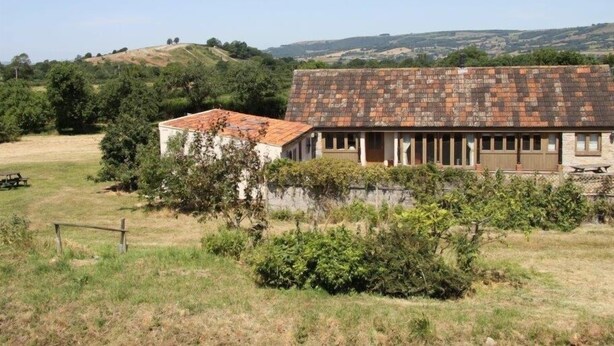 A rustic building with a tiled roof, surrounded by greenery and hills in the background. - The Milking Parlour (Wells)