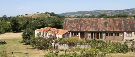 Un bâtiment rustique avec un toit en tuiles, entouré de verdure et avec des collines en arrière-plan.