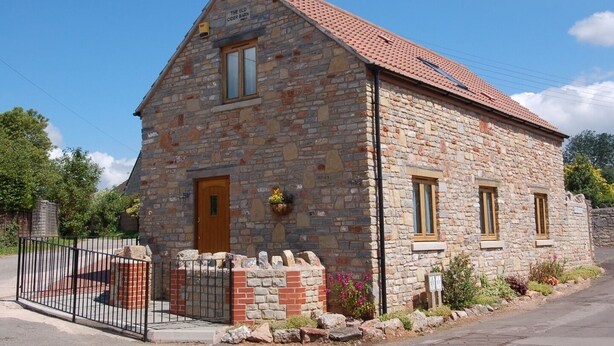 A stone building with a tiled roof, a wooden door, and a small garden with flowers. - The Old Cider Barn (Wells)