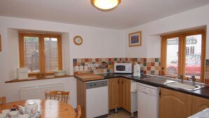 A compact kitchen with wooden cabinets, a microwave, a sink, and a dining table set with tea cups and saucers. - The Old Cider Barn (Wells)
