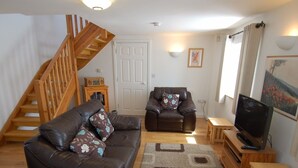 A living room with a wooden staircase, a brown leather sofa, a brown leather armchair, a television, and a wooden coffee table. - The Old Cider Barn (Wells)