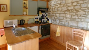 A compact kitchen with wooden cabinets, a stone wall, a microwave, a kettle, and a sink.