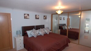 A bedroom with a large bed, a nightstand, a lamp, a mirror, and a ceiling light. - Garden View (Newton Abbot)