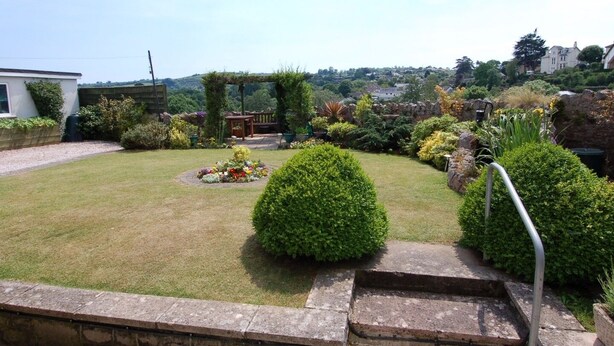 A well-maintained garden with a lawn, various shrubs, a flower bed, and a stone pathway leading to a wooden bench. - Garden View (Newton Abbot)