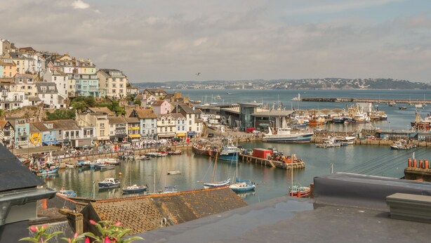 Une ville côtière aux bâtiments aux couleurs vives, un port de plaisance rempli de bateaux et un phare visible au loin.
