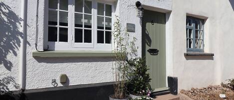 A white building with a green door, a window with white shutters, and a small porch area with potted plants.