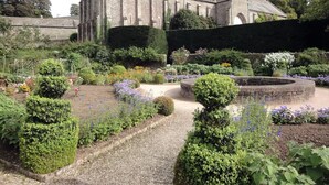 A historic building with a symmetrical facade, surrounded by a well-maintained garden with various flowering plants and neatly trimmed hedges. - Burham Barn (Yelverton)