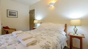 A neatly made bed with a floral bedspread and folded towels, two bedside lamps, a wooden nightstand, a wicker chair with a red cushion, and a framed picture on the wall.