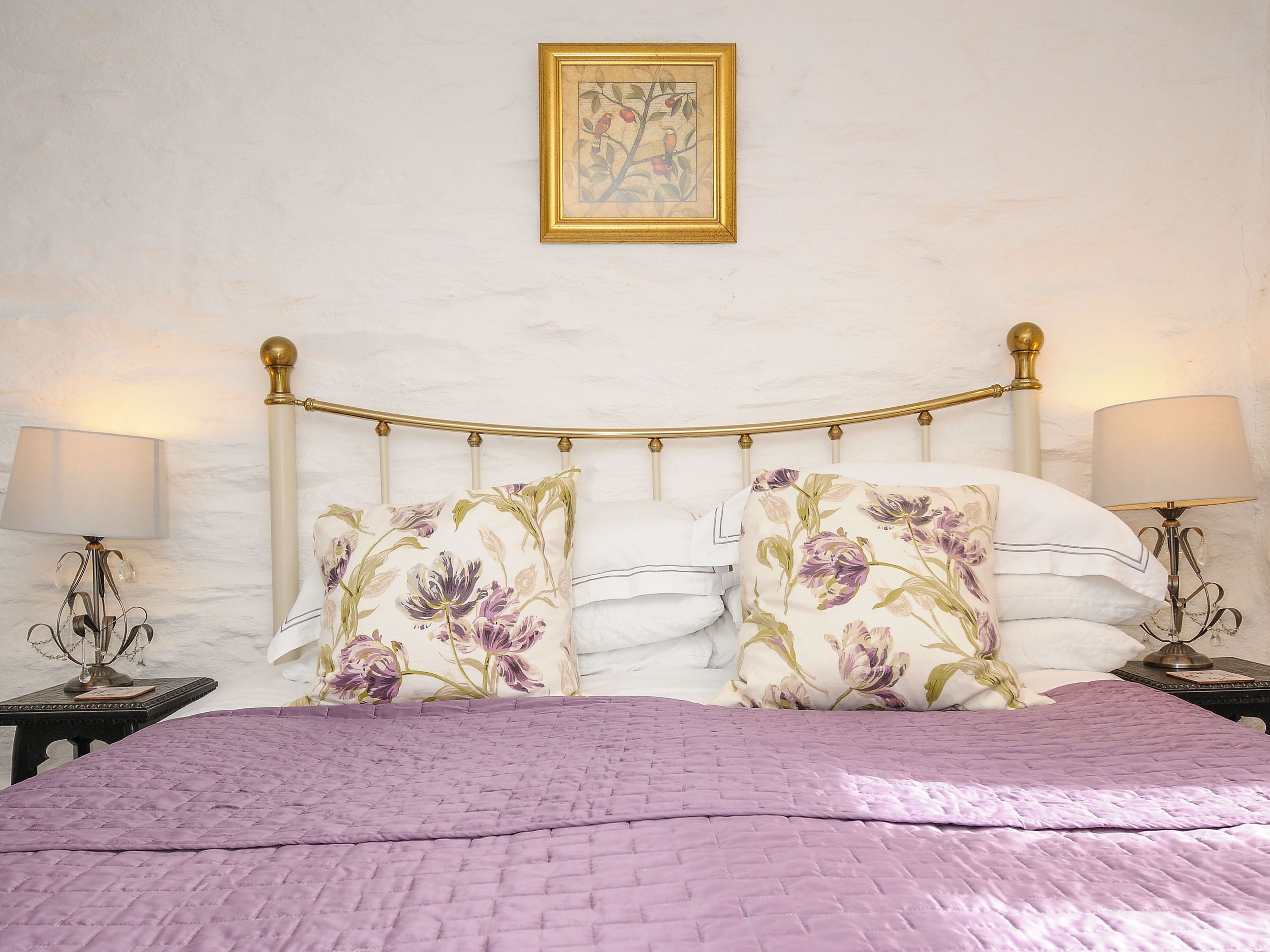 A bedroom with a bed, floral pillows, a headboard, two bedside tables with lamps, and a framed picture on the wall.