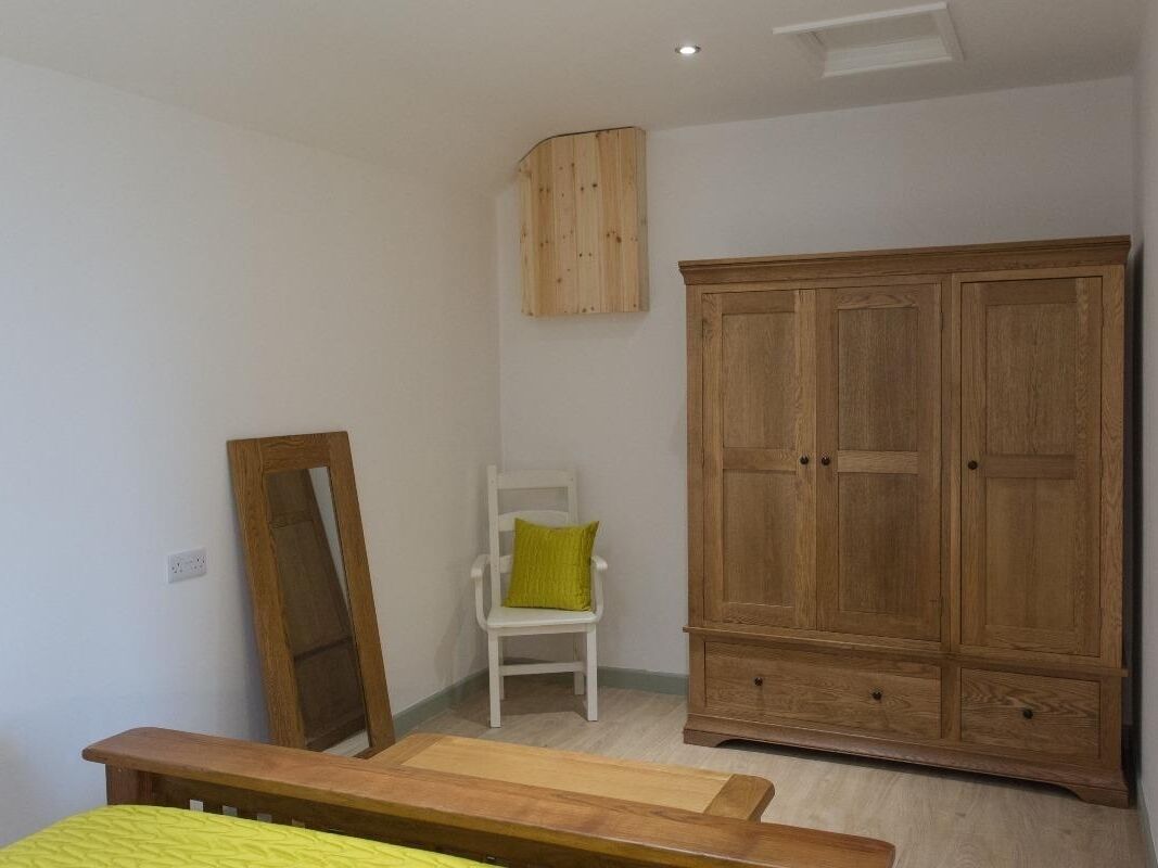 A bedroom with a wooden wardrobe, a white chair with a green cushion, and a wooden bench.