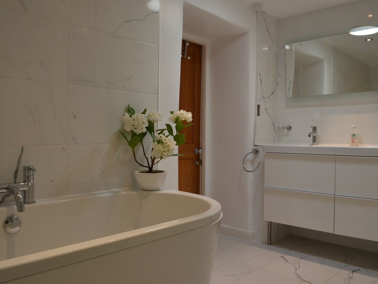 Un baño con una bañera independiente, un lavamanos con espejo y una planta en maceta con flores blancas.