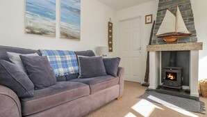 A living room with a grey sofa, a fireplace with a stone mantle, and two paintings on the wall. - Cockle Island Cottage (Helston)