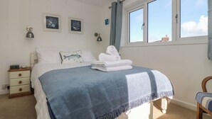 A bedroom with a bed, a nightstand, a chair, and a window with curtains. - Cockle Island Cottage (Helston)