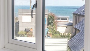 A window view of a residential area with a clear sky, a body of water, and houses with visible roofs and fences. - Cockle Island Cottage (Helston)