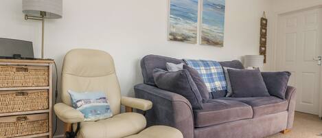 A living room with a beige armchair, a grey sofa, and a small wooden side table. There are two wall-mounted paintings and a lamp on the side table.
