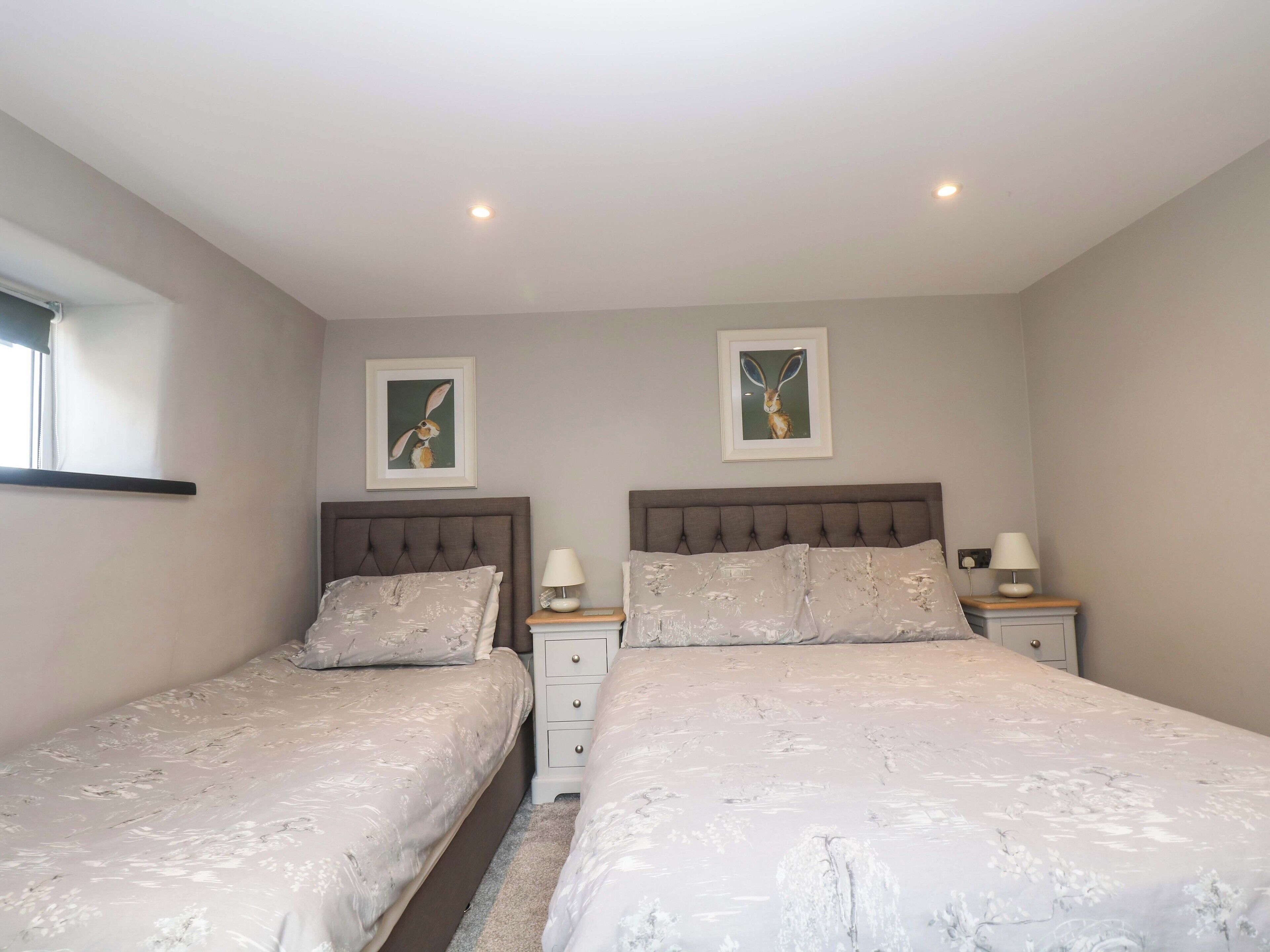 A bedroom with two beds, each with a headboard and nightstands. There are two framed pictures on the wall, a window on the left, and a ceiling light.