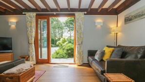 A cozy living room with a wooden ceiling, a sofa with cushions, a television, and a window with curtains. - Oaklands (Camelford)