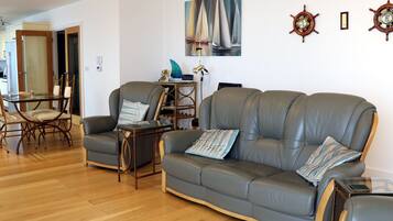 Een woonkamer met een grijze leren bank, een glazen salontafel en een eethoek met een glazen tafel en stoelen.